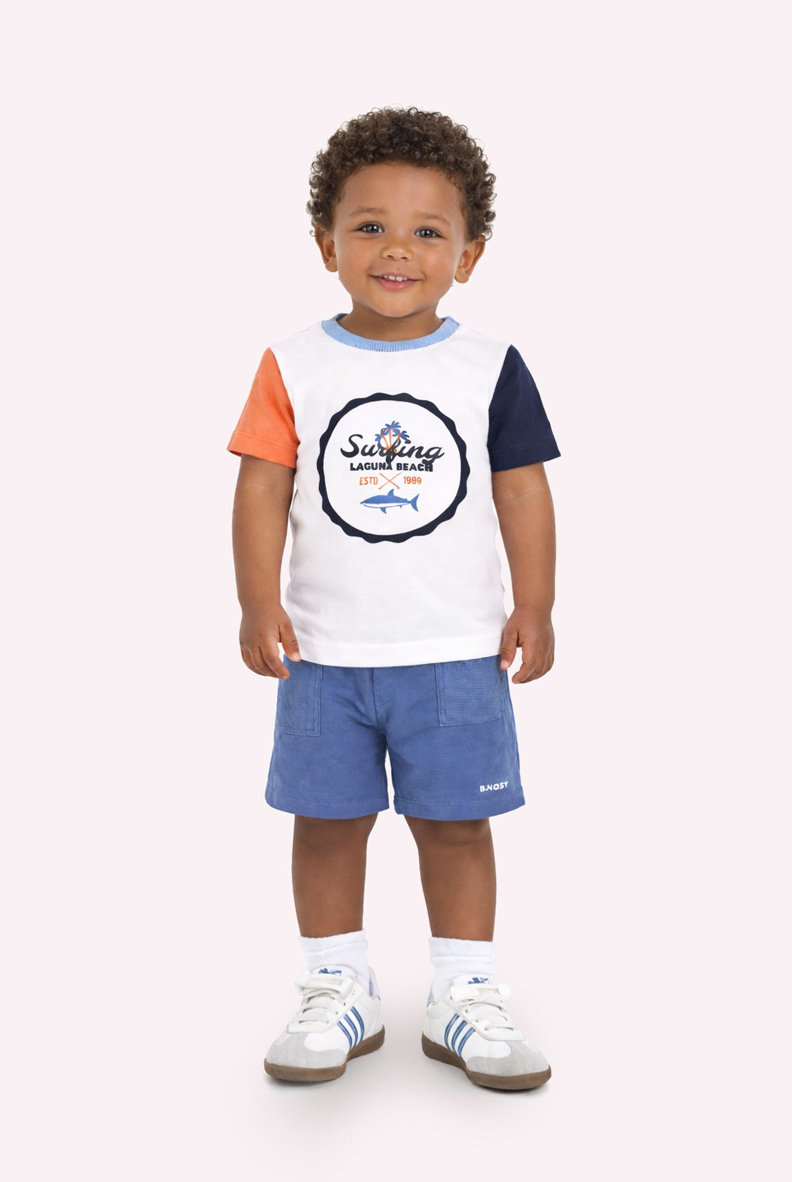 Een jonge jongen staat lachend in een stijlvolle, kleurrijke t-shirt en korte broek, met een warme achtergrond.